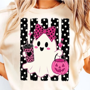 Op de afbeelding: Een crèmekleurig t-shirt met een afbeelding van een witte geest met een roze strik met stippen, die een roze beker met luipaardprint en een roze pompoenemmer vasthoudt. De achtergrond bestaat uit zwarte verticale strepen met witte stippen.