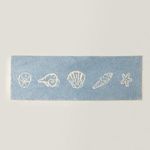 May include: A rectangular, light blue bath mat featuring five white seashell designs. The seashells include a spiral, a conch, a scallop, a spiral shell, and a starfish. The mat is made of a soft, textured material.