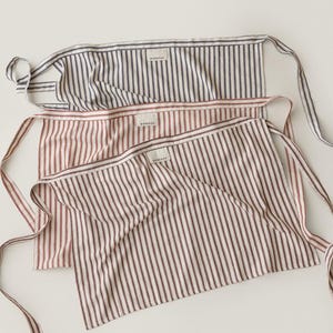 May include: Three striped aprons in varying colors: navy and white, red and white, and a darker red and white. Each apron has a rectangular body with vertical stripes and long ties. The aprons are made of a soft fabric and have a tag that reads "WEEKEND".