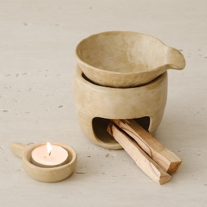 May include: A beige ceramic essential oil diffuser with a small bowl on top and a cavity for incense sticks. A lit tea light candle sits in a matching small dish. Palo santo sticks are placed in the diffuser.