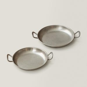May include: Two small, round, silver-toned metal pans with loop handles. The pans have a shallow depth and a slightly textured surface. The handles are attached to the sides of the pans, providing a secure grip. The pans are likely used for cooking or serving.