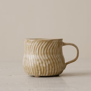 May include: A beige ceramic mug with a textured, wavy pattern. The mug has a rounded base, a curved handle, and a smooth rim. The mug is a neutral color and appears to be handmade.