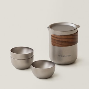 May include: A stainless steel tea set with a wooden handle and the word "WENSHUO" printed on the side. The set includes a teapot, a lid, and three small, round tea bowls. The tea set is designed for brewing and serving tea.