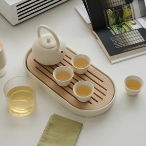 May include: A serene tea set arrangement featuring a white teapot, small cups, and a wooden tray. A glass of tea and a green napkin are also present. A white record player and a book are in the background, creating a calming aesthetic.