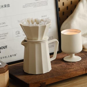May include: A cream-colored ceramic pour-over coffee maker with a paper filter, a small candle, and a wooden tray. The coffee maker has a geometric design. The text on the poster says "Three legs Fruit Plate".