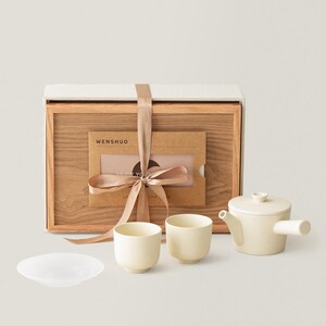 May include: A cream-colored tea set with a teapot, two cups, and a small plate. The set is presented with a wooden box tied with a ribbon. The box contains a card with the text "Wenshuo".