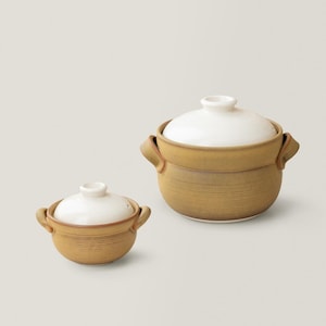 May include: Two tan ceramic cooking pots with white lids. The pots have a round shape with handles on the sides. The lids have a small knob on top. The pots are of different sizes, with the larger one on the right.