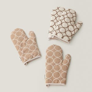 May include: Three tan oven mitts and pot holders with a white dotted circle pattern. The mitts and pot holders have a white trim and are arranged on a light background.