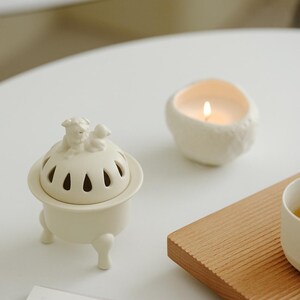 May include: Off-white ceramic incense burner with a dog figurine on top, featuring a lid with teardrop-shaped cutouts. A lit candle in a textured, off-white holder and a wooden tray are also present, creating a serene, minimalist aesthetic.