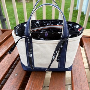 May include: A cream-colored canvas tote bag with navy blue trim and two handles. The interior features a black fabric lining with a repeating white skeleton pattern. Two black fabric ties are attached to the front handles, and a zippered pocket is visible inside the main compartment.