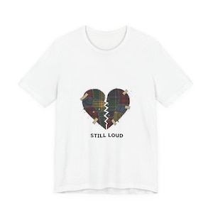 Puede incluir: Camiseta blanca con un diseño de corazón roto. El corazón está dividido, con un patrón de retazos en tonos verdes, rojos y amarillos. El texto "STILL LOUD" está impreso debajo.