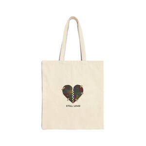 May include: A cream-coloured tote bag with a heart design. The heart is split, patched with tape, and has a plaid pattern. The text "STILL LOUD" is printed below. The bag has two handles and is made of a canvas-like material.