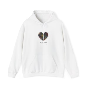 Puede incluir: Sudadera con capucha blanca con bolsillo delantero y capucha con cordón. El diseño presenta un gráfico de corazón roto en cuadros con el texto "STILL LOUD" debajo. La sudadera con capucha es una prenda informal.