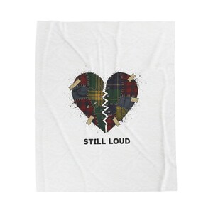 May include: White blanket featuring a heart design with a broken center. The heart is composed of patchwork plaid patterns in red, green, and blue. The text "STILL LOUD" is printed below the heart.
