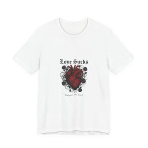 Puede incluir: Camiseta blanca con un diseño de corazón anatómico rojo oscuro. El texto "Love Sucks" está encima del corazón, con "Embrace the Dark" debajo. El diseño incluye acentos de rosas negras.
