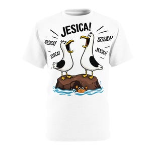Puede incluir: Camiseta blanca con una ilustración de dibujos animados de dos gaviotas en una roca, gritando "JESICA!". El diseño incluye el texto "JESSICA!" y un pequeño cangrejo naranja en el agua. Una camiseta gráfica humorística.