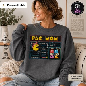 Puede incluir: Sudadera gris oscuro de Pac Mom con un diseño de Pac-Man. El diseño incluye un personaje amarillo de Pac-Man con un lazo rojo, fantasmas y los nombres Liam, Emma, Noah y Ava. La sudadera es unisex y de mezcla de algodón suave.
