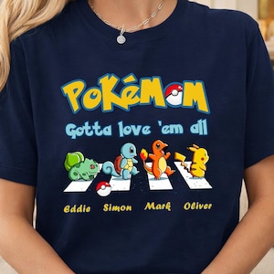 Puede incluir: Camiseta azul marino con la palabra "Pokémon" en amarillo y rojo, con la frase "Gotta love 'em all". Cuatro personajes de Pokémon cruzan una carretera con rayas blancas. Los nombres "Eddie", "Simon", "Mark" y "Oliver" están debajo.