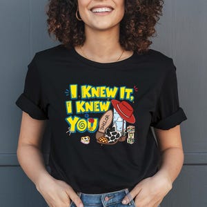 K&ouml;nnte beinhalten: Ein schwarzes Kurzarm-T-Shirt mit einem Grafikdruck und dem gelben Text I KNEW IT, I KNEW YOU. Das Design zeigt einen roten Cowboyhut, einen wei&szlig;-schwarzen Cowboystiefel mit Kuhfleckenmuster und der Aufschrift TAYLOR sowie bunte Baukl&ouml;tze.