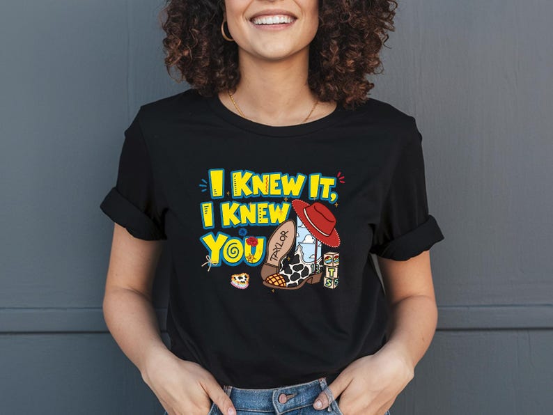 K&ouml;nnte beinhalten: Ein schwarzes Kurzarm-T-Shirt mit einem Grafikdruck und dem gelben Text I KNEW IT, I KNEW YOU. Das Design zeigt einen roten Cowboyhut, einen wei&szlig;-schwarzen Cowboystiefel mit Kuhfleckenmuster und der Aufschrift TAYLOR sowie bunte Baukl&ouml;tze.