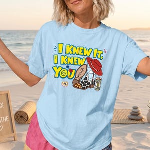K&ouml;nnte beinhalten: Ein hellblaues T-Shirt mit kurzen &Auml;rmeln und einem Grafikdruck mit dem gelben Text I KNEW IT, I KNEW YOU. Das Design zeigt einen roten Cowboyhut, einen Stiefel mit Kuhfleckenmuster und dem Namen TAYLOR, eine kleine Blume und drei bunte W&uuml;rfel.