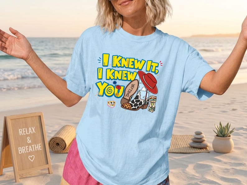 K&ouml;nnte beinhalten: Ein hellblaues T-Shirt mit kurzen &Auml;rmeln und einem Grafikdruck mit dem gelben Text I KNEW IT, I KNEW YOU. Das Design zeigt einen roten Cowboyhut, einen Stiefel mit Kuhfleckenmuster und dem Namen TAYLOR, eine kleine Blume und drei bunte W&uuml;rfel.