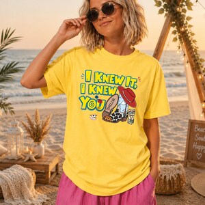 K&ouml;nnte beinhalten: Ein leuchtend gelbes T-Shirt mit dem Text I KNEW IT, I KNEW YOU in blauen und gelben Buchstaben. Die Grafik zeigt einen Cowboystiefel mit Kuhmuster, einen roten Hut, eine kleine Blume und einen Spielzeugblock. Das T-Shirt hat kurze &Auml;rmel.