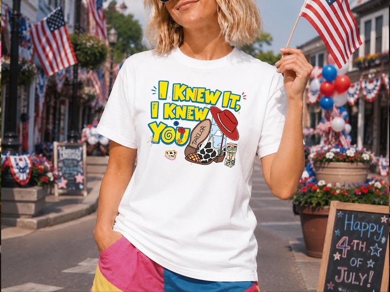 K&ouml;nnte beinhalten: Ein wei&szlig;es Kurzarm-T-Shirt mit einem Grafikdruck und dem gelben Text I KNEW IT, I KNEW YOU. Das Design zeigt einen Cowboystiefel mit Kuhfellmuster und dem Namen TAYLOR, einen roten Cowboyhut sowie bunte Bausteine mit aufgedruckten Buchstaben.