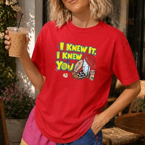 K&ouml;nnte beinhalten: Ein leuchtend rotes T-Shirt mit kurzen &Auml;rmeln und einem Grafikdruck mit dem Text I KNEW IT, I KNEW YOU in Gelb und Blau. Das Design zeigt einen Cowboystiefel mit Kuhfleckenmuster, einen roten Hut und einen kleinen Block mit den Buchstaben TS.
