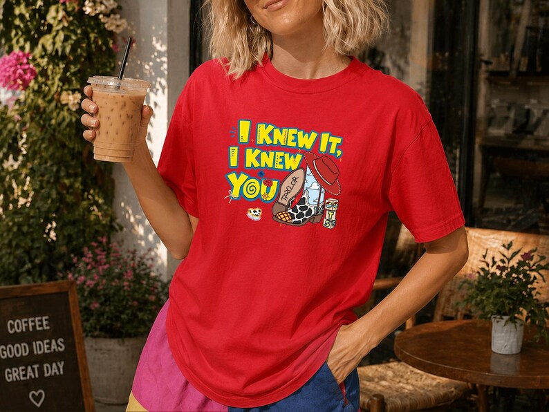 K&ouml;nnte beinhalten: Ein leuchtend rotes T-Shirt mit kurzen &Auml;rmeln und einem Grafikdruck mit dem Text I KNEW IT, I KNEW YOU in Gelb und Blau. Das Design zeigt einen Cowboystiefel mit Kuhfleckenmuster, einen roten Hut und einen kleinen Block mit den Buchstaben TS.