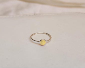 May include: A delicate silver ring with a small, round, pale yellow stone set in the center. The ring band is thin and polished, and the stone has a subtle, matte finish. The ring is displayed on a light-colored surface.