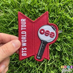May include: A red and white embroidered patch featuring the text "BTS WORLD TOUR" and a stylized light stick design. The light stick is white with red accents and a black handle. The patch is held against a background of green grass.