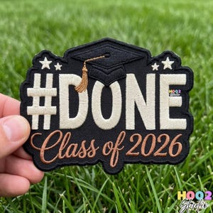 May include: Black and white embroidered patch with the word "DONE" and "Class of 2026". Features a graduation cap and two stars. The patch is held against a background of green grass.