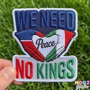 May include: Embroidered patch with the words "WE NEED" in navy blue, above a heart-shaped design formed by hands. The heart says "Peace" and is surrounded by red, white, and green colors. Below, the patch reads "NO KINGS" in red and green.