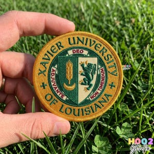 May include: A round, embroidered patch with the words "Xavier University of Louisiana" in a gold border. The patch features a green and gold crest with a lion, wheat, and crescent moon design. The patch is held in a person's hand.