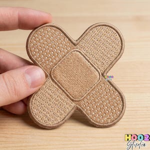 May include: A tan-colored embroidered patch in the shape of a bandage. The patch features a textured design with a central rectangular pad and four rounded flaps. The patch is held up by a hand. The text "HOOZ Studio" is visible.
