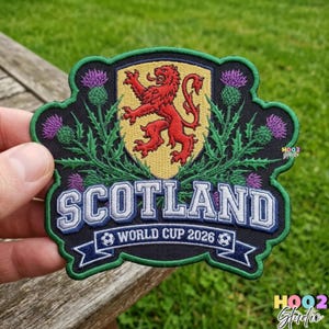 May include: Embroidered patch featuring the Scotland national emblem, a red lion rampant on a gold shield, surrounded by purple thistles and green foliage. The patch reads "SCOTLAND WORLD CUP 2026" in white and blue.