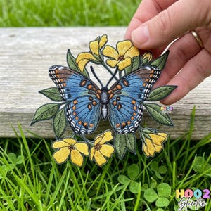 May include: Embroidered patch featuring a blue butterfly with orange and brown details, surrounded by yellow flowers and green leaves. The patch is held against a wooden surface and green grass.