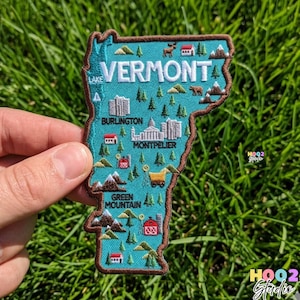 May include: Embroidered patch in the shape of Vermont, with teal background and brown border. The patch features the word "VERMONT" and illustrations of landmarks, mountains, and trees. The cities of Burlington and Montpelier are also labeled.