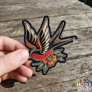 May include: Embroidered patch featuring a traditional swallow bird design. The bird has black and tan wings, a red breast, and is perched above an orange flower with green leaves. The patch has a black border.