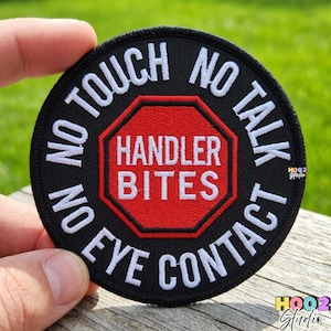 May include: A round black patch with white embroidered text that reads "NO TOUCH, NO TALK, NO EYE CONTACT." The center features a red octagon with "HANDLER BITES" in white. The patch is held by a hand.