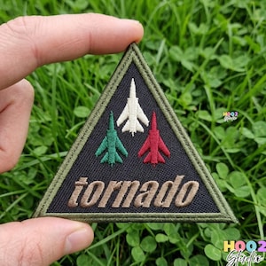 May include: A triangular patch with a black background and a green border. It features three embroidered jet plane designs in green, white, and red. The word "tornado" is embroidered in tan. The patch is held against a backdrop of green grass.