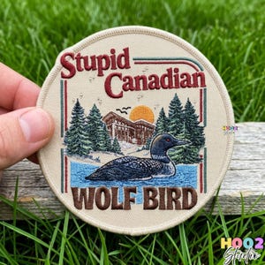 May include: Round embroidered patch with the text "Stupid Canadian" and "WOLF BIRD". The patch features a loon swimming in water, a cabin, pine trees, and a setting sun. The patch is beige with brown, red, green, and blue accents.
