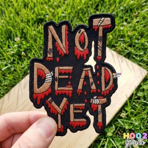 May include: Black embroidered patch with the words "NOT DEAD YET" in a distressed, bloody font. The patch features brown and red accents, with a hammer graphic. The patch is held against a background of green grass.