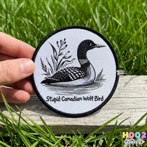 May include: Round, black and white patch featuring a stylized illustration of a loon swimming in water, with aquatic plants. The text "Stupid Canadian Wolf Bird" is printed below the image. The patch has a black border.
