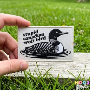 May include: A rectangular patch featuring a black and white illustration of a loon bird on water. The text "stupid canadian wolf bird" is above the bird. The patch is held against a background of green grass.