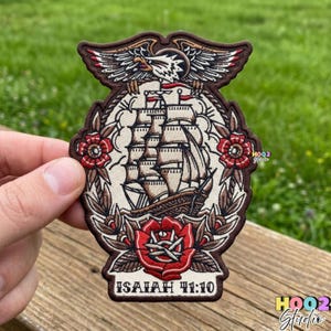 May include: Embroidered patch featuring a detailed illustration of a tall ship, an eagle, and roses. The patch has a brown border and includes the text "ISAIAH 41:10". The design is in shades of brown, red, and cream.