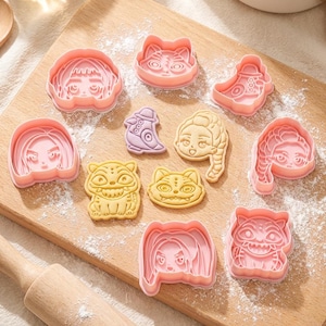 May include: A collection of pink plastic cookie cutters in various shapes, including faces, a cat, and a boot, arranged on a wooden cutting board dusted with flour. A wooden rolling pin is visible in the lower left corner.