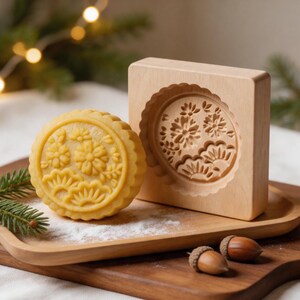 Könnte beinhalten: Ein runder, goldgelber Mondkuchen mit einem detaillierten Blumen- und Kiefernnadelmuster, platziert neben einer quadratischen Holzform mit demselben komplizierten Prägedesign. Die Form besteht aus hellem Holz mit einer kreisförmigen Schnitzerei.