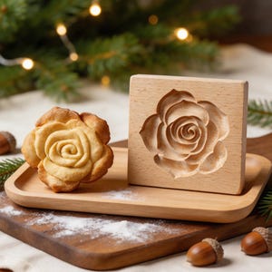 Könnte beinhalten: Eine Holzkeksform und ein rosenförmiger Keks liegen auf einem Holztablett. Die Form ist quadratisch mit einem geschnitzten Rosendesign. Der gebackene Keks ist goldbraun und ähnelt dem Design der Form. Die Szene spielt auf einem Holzbrett mit festlichem Grün.